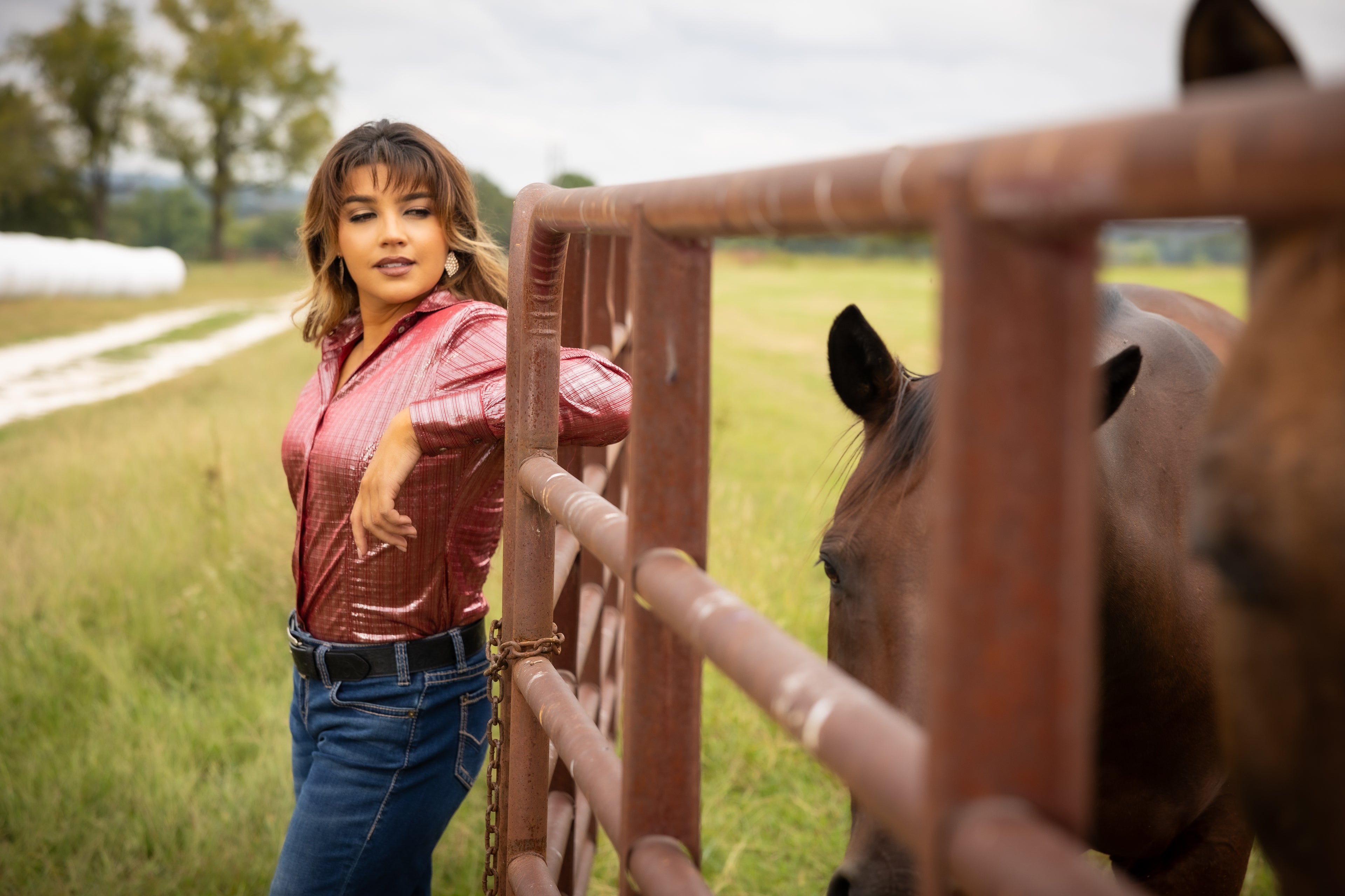 Model wearing Ruby shirt from Kultured Rider's jewel collection, posing in ranch setting with horse 